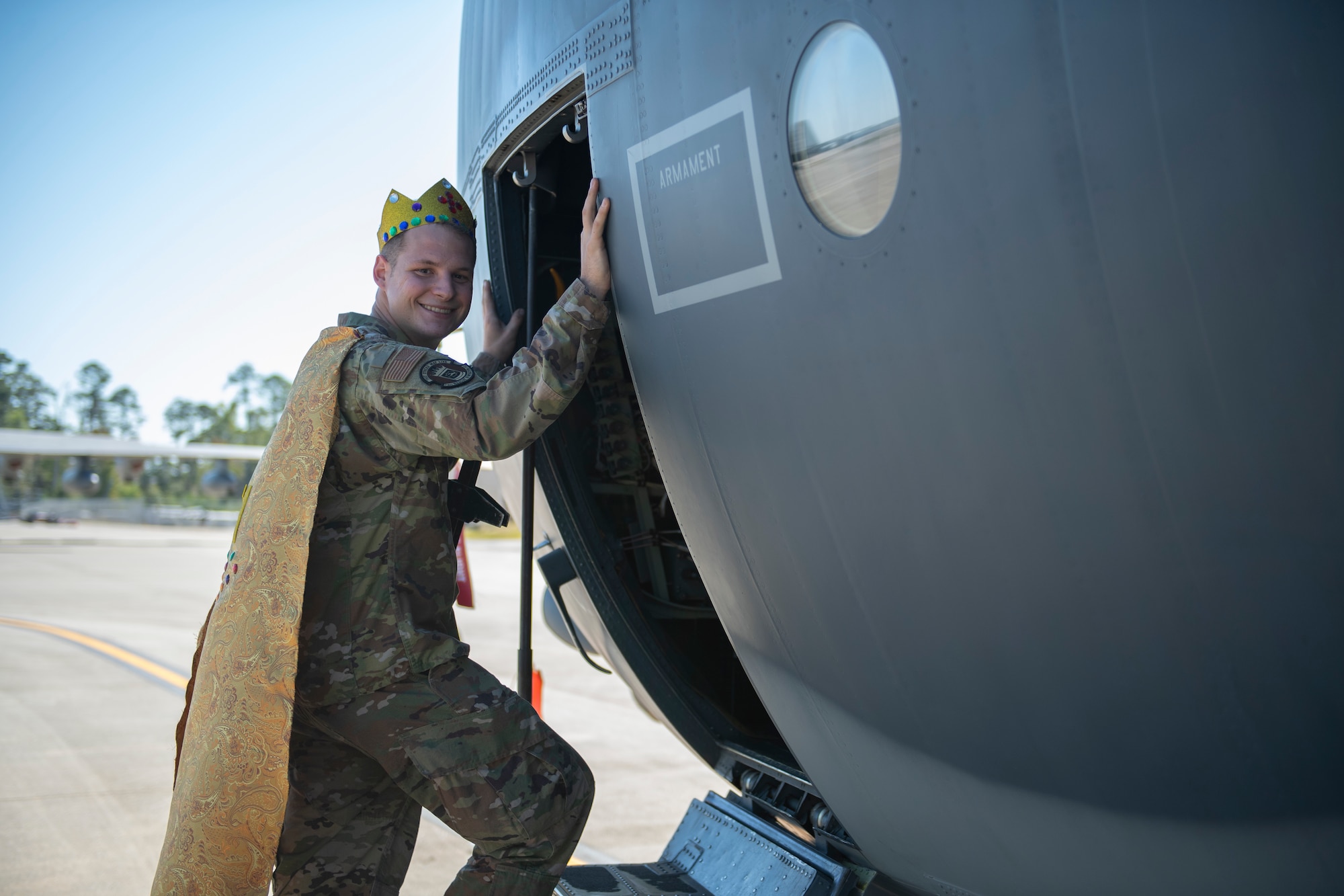 A photo of a man stepping inside an HC-130J, posing for a photo wearing a gold and bedazzled crown and cape.