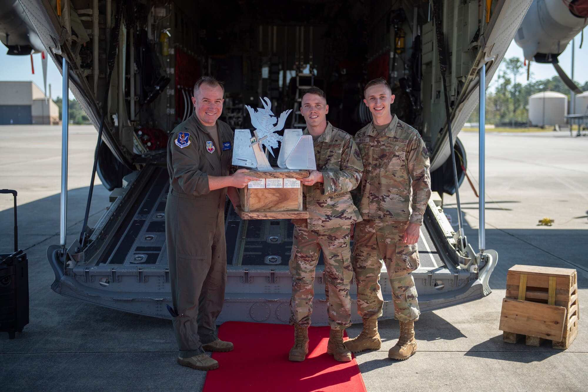 A photo of three people holding a big metal trophy. They are standing on a red carpet laid out behind an HC-130J with its door down/open.