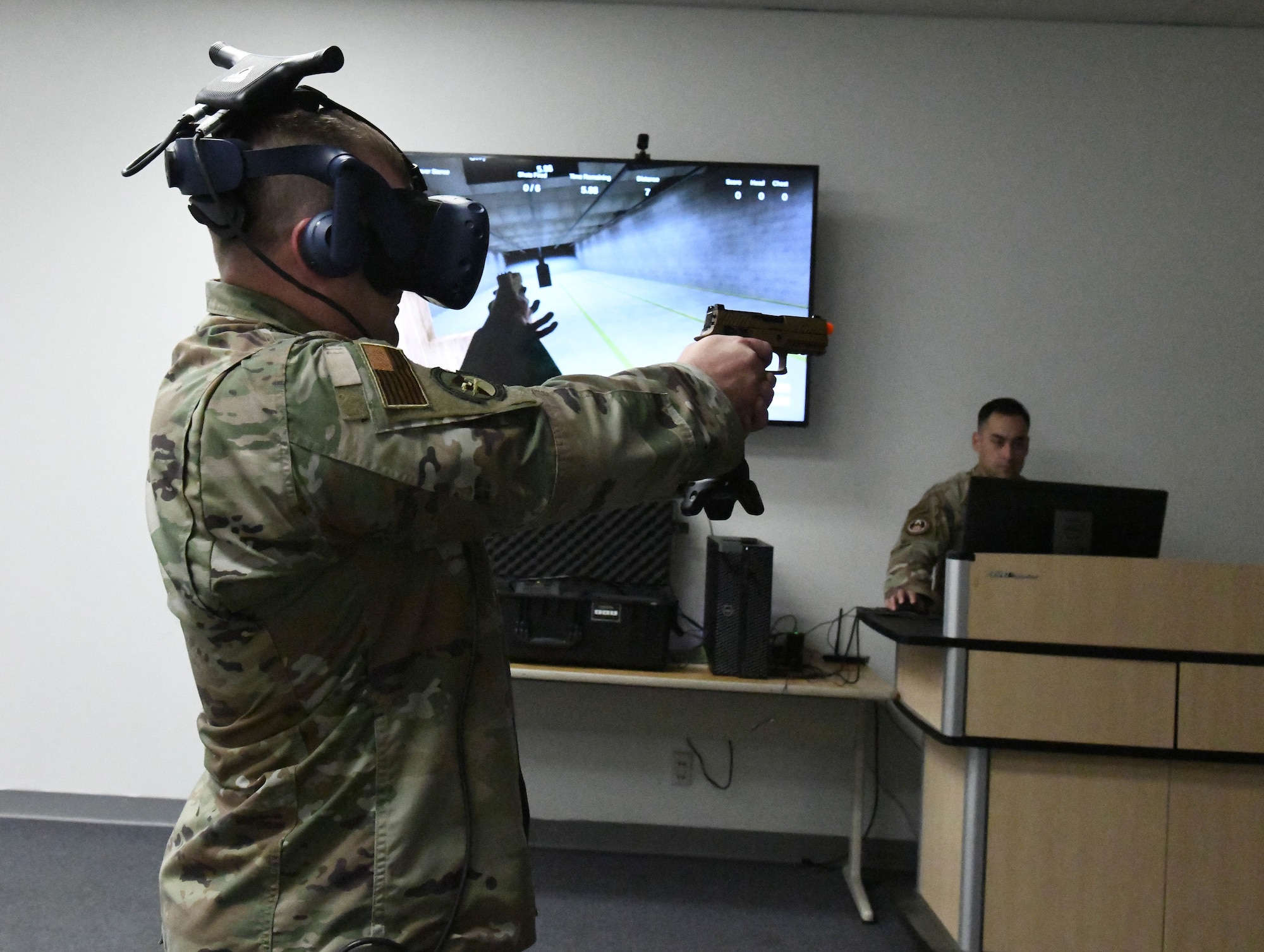 An airman wearing a virtual realty headset holds a gun simulator as another airman runs a simulator training.