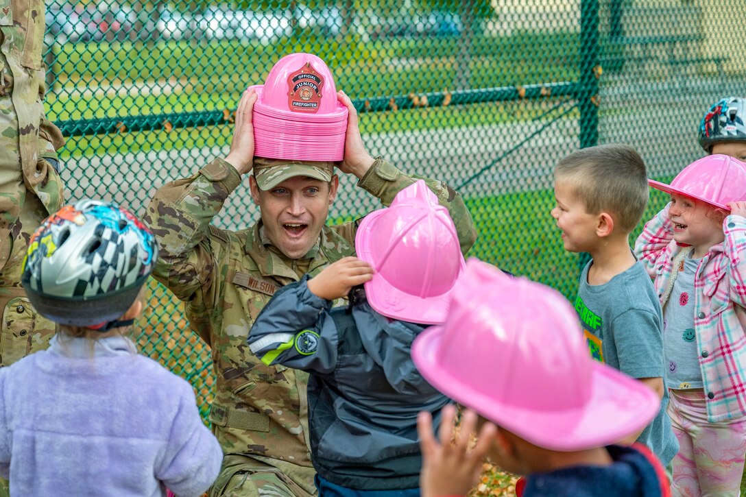 An airman holds a stack of pink plastic hats on his head as children watch.