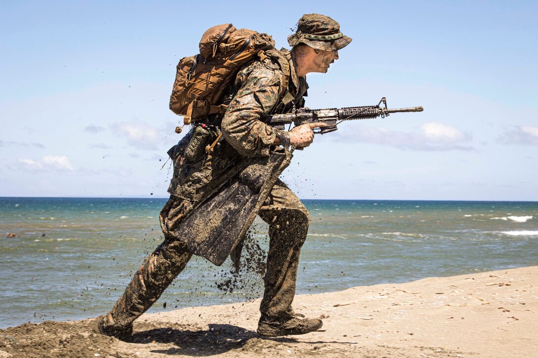 A Marine covered in sand aims a weapon while walking on the beach.