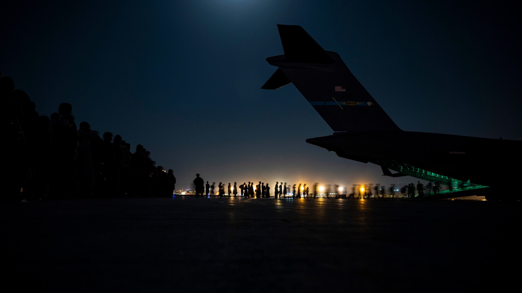 People line up to walk on to a large military aircraft. The photo was taken at night, only silhouettes of people are visible.  