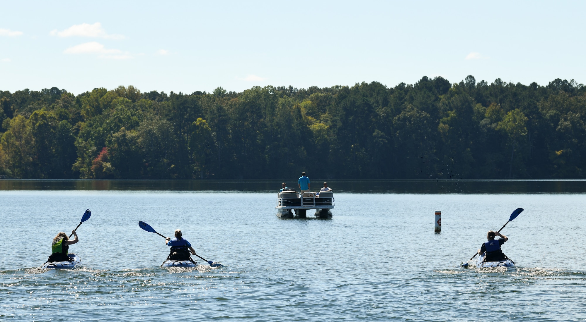 Three people paddle kayaks toward a pontoon boat
