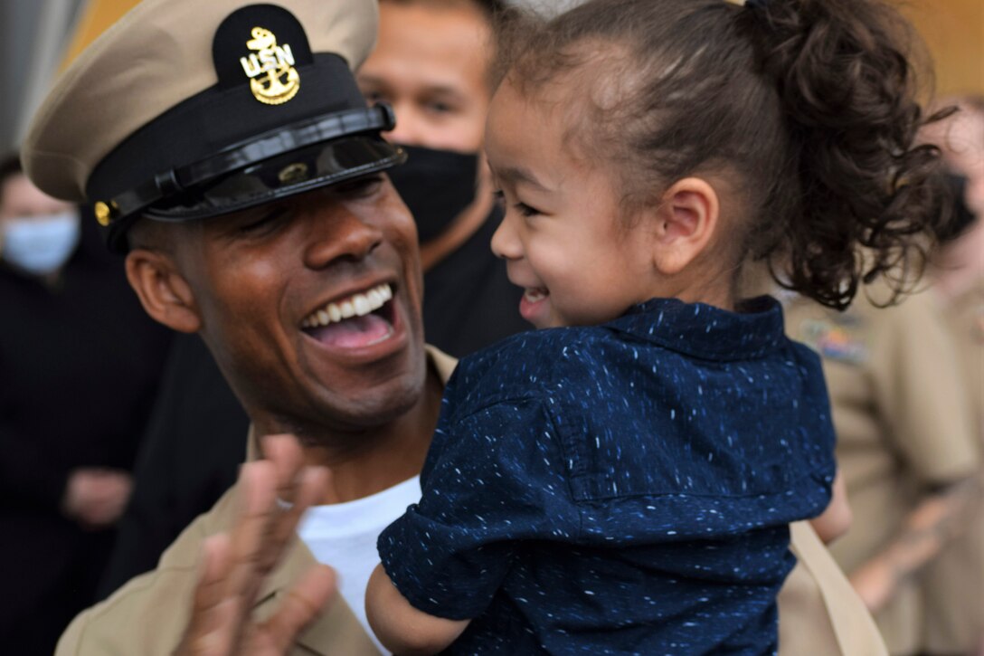 A sailor laughs with his daughter with a crowd in the background.