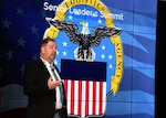 Mr. Cannon stands on a stage with the DLA logo behind him