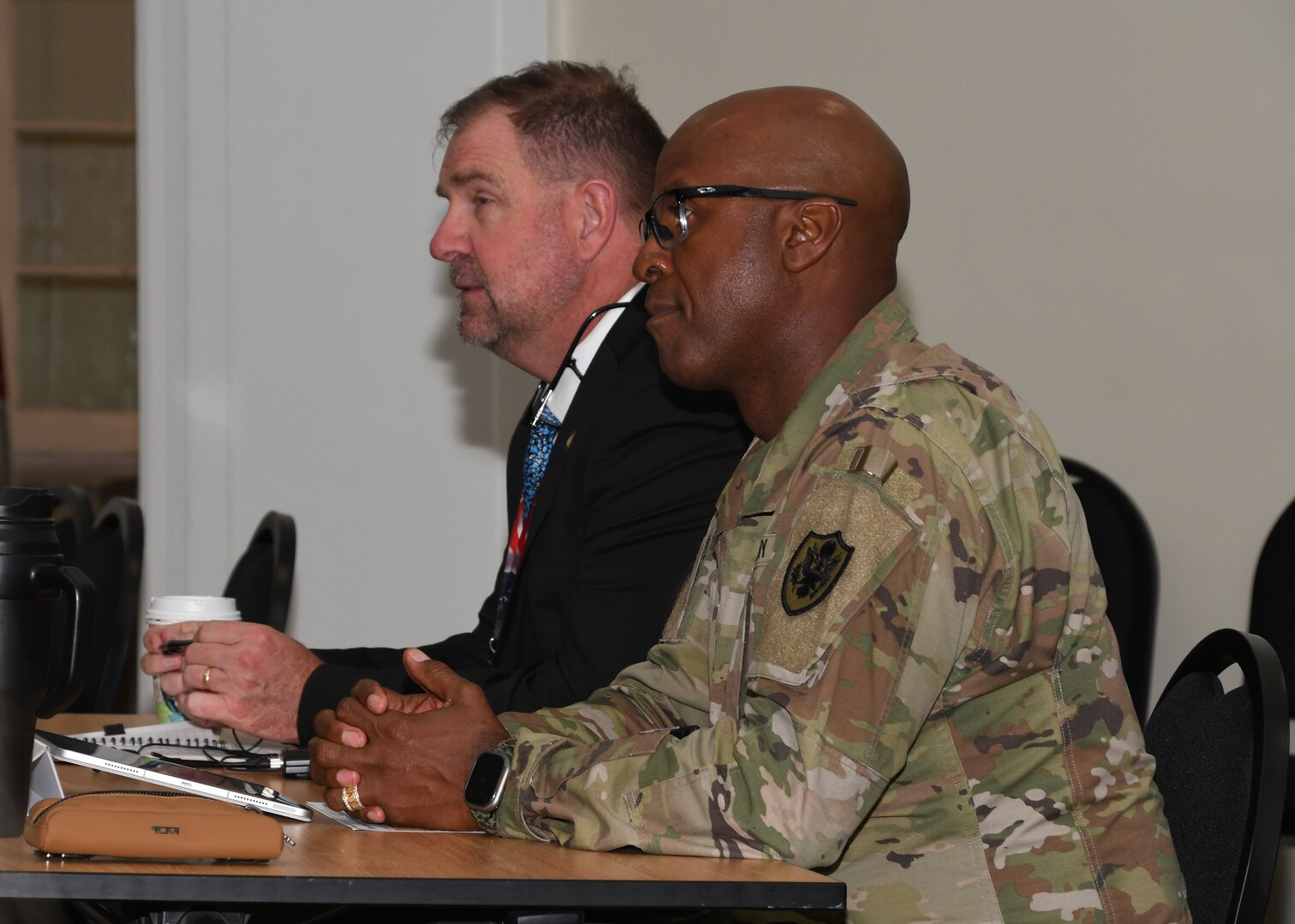 Defense Logistics Agency Disposition Services Director Mike Cannon in a civilian suite and U.S. Army Col. Andre L. Toussaint DLA Disposition Services deputy director, in his Army uniform, sit at a table and listen to a conversation at their command’s Senior Leader Summit held in Battle Creek, Michigan.