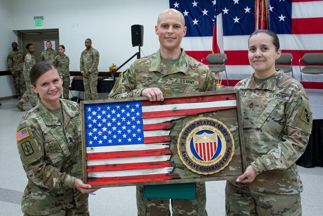First Sgt. Leesa Petty, outgoing first sergeant, and Sgt. 1st Class Marina Carrillo, senior human resources non-commissioned officer, Headquarters and Headquarters Detachment, 75th Innovation Command, present outgoing HHD commander Maj. Christopher Allen a custom plaque as a token of the command's appreciation of Allen's three-year tenure. The wooden flag was hand-constructed by Col. Steven Waldrop, the 75th Innovation Command's G3/5/7 chief. (U.S. Army photo by Maj. Charles An, 75th Innovation Command)