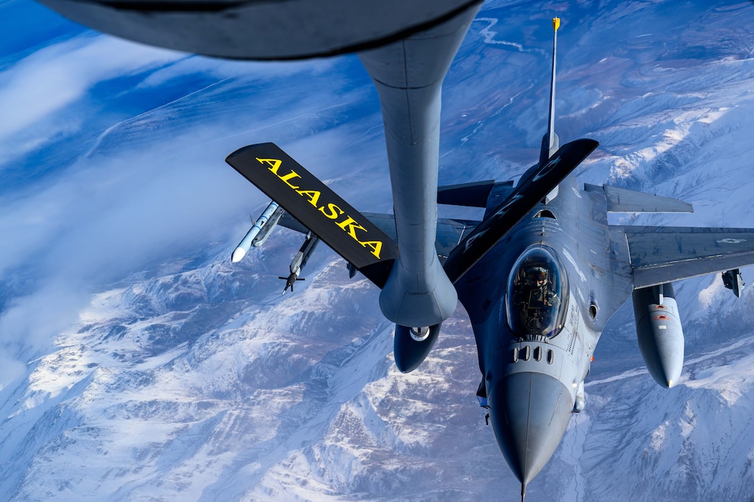 A military aircraft prepares to be refueled as it flies above snowy mountains.