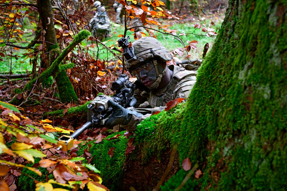 A soldier in camouflage lies on forest floor by a tree stump pointing a weapon.