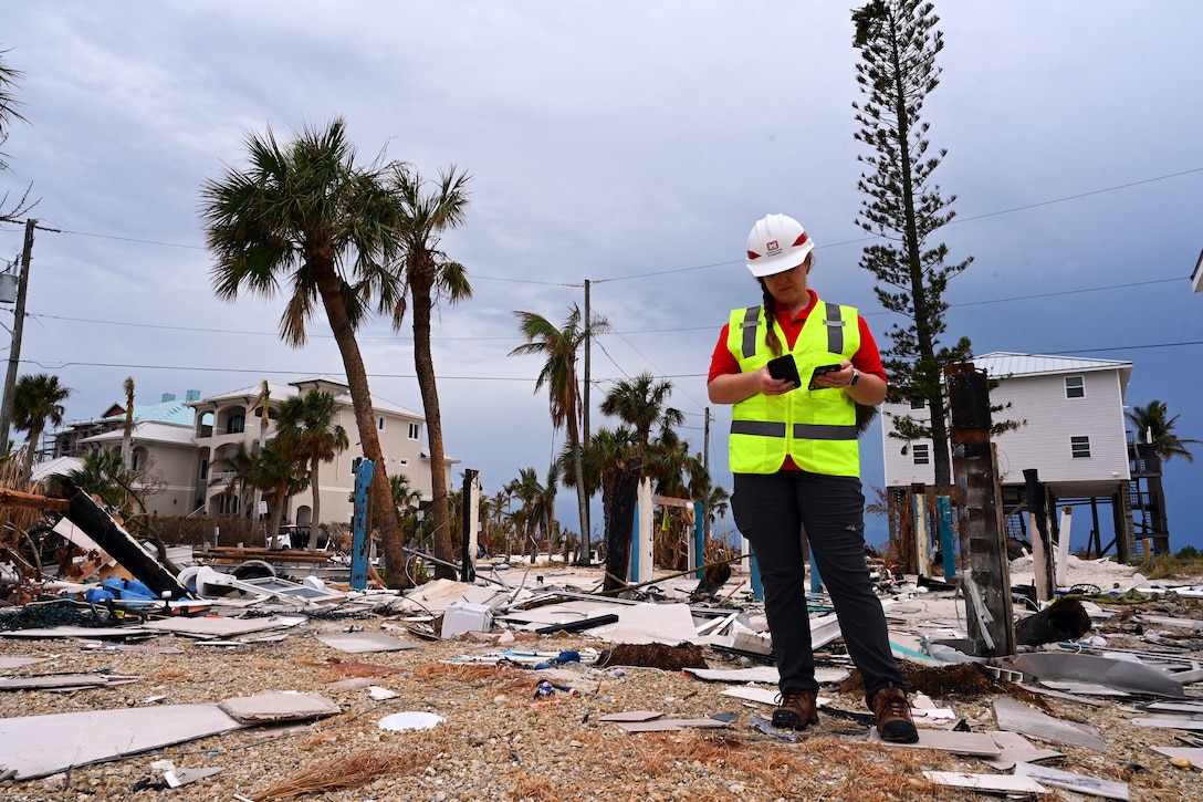 An Army Corps of Engineers worker in neon vest and hardhat holds two phones while standing on damaged beach.