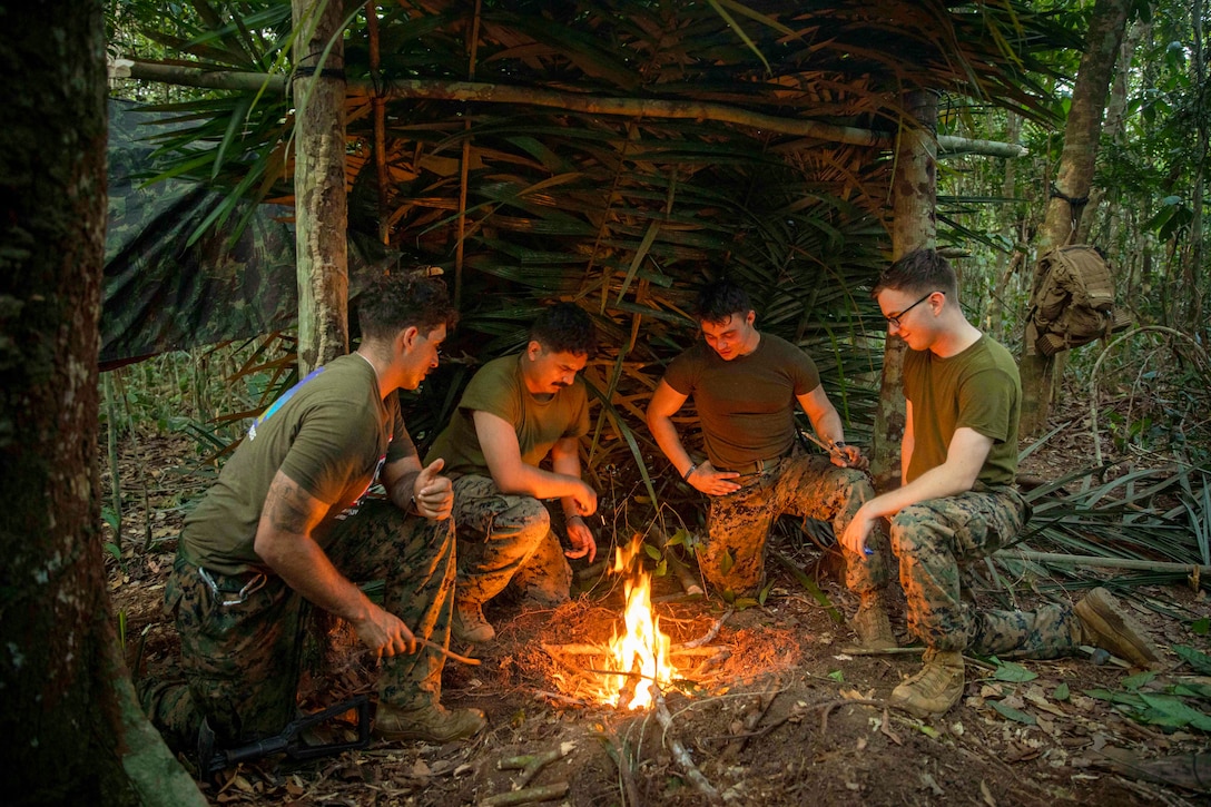 Marines and sailors gather around a fire in the woods.