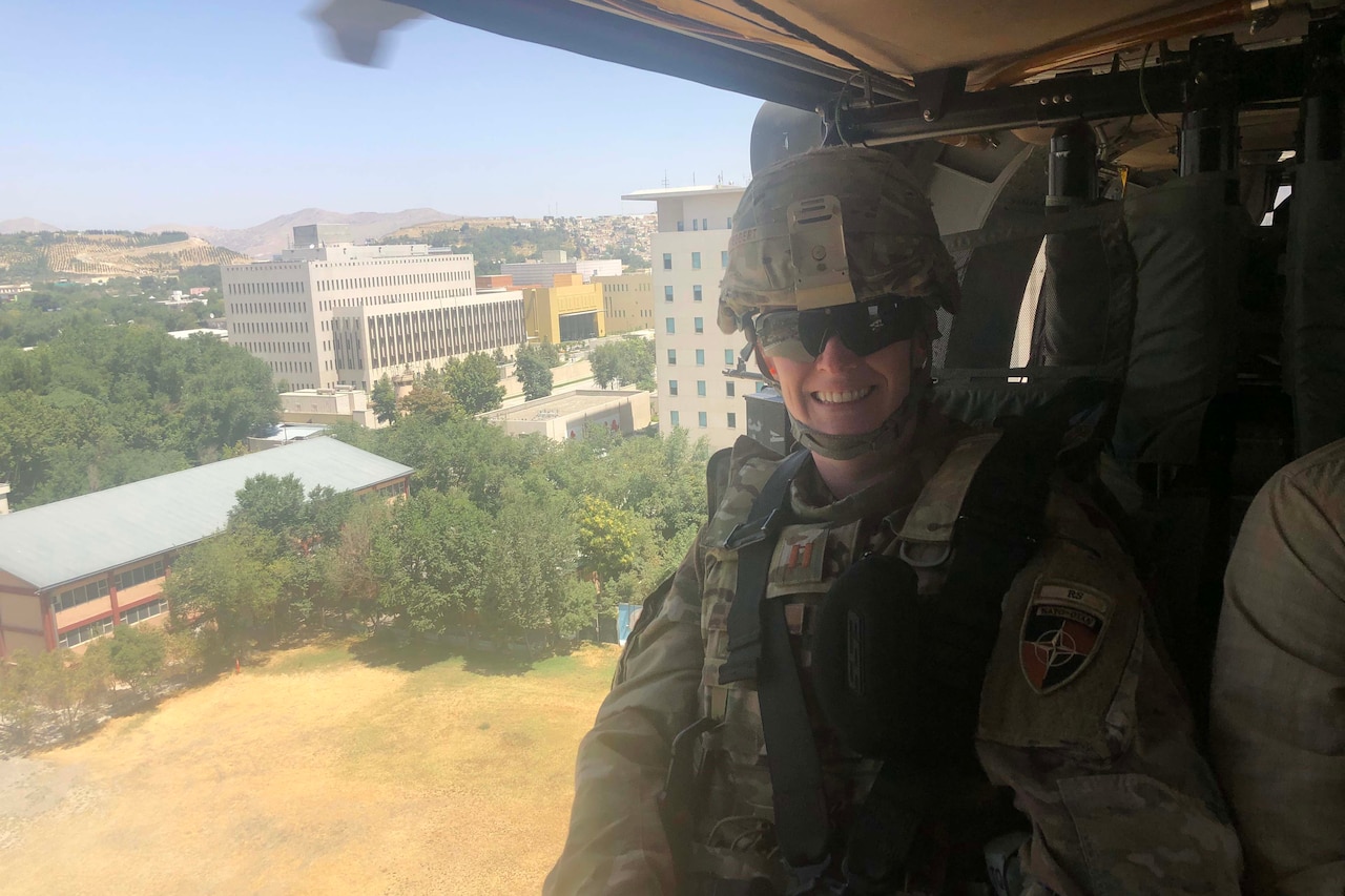 A woman in combat uniform smiles from inside a helicopter as it flies over a desert town.