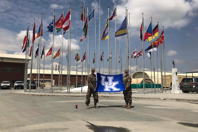 Two people hold up a flag in front of several flagpoles.