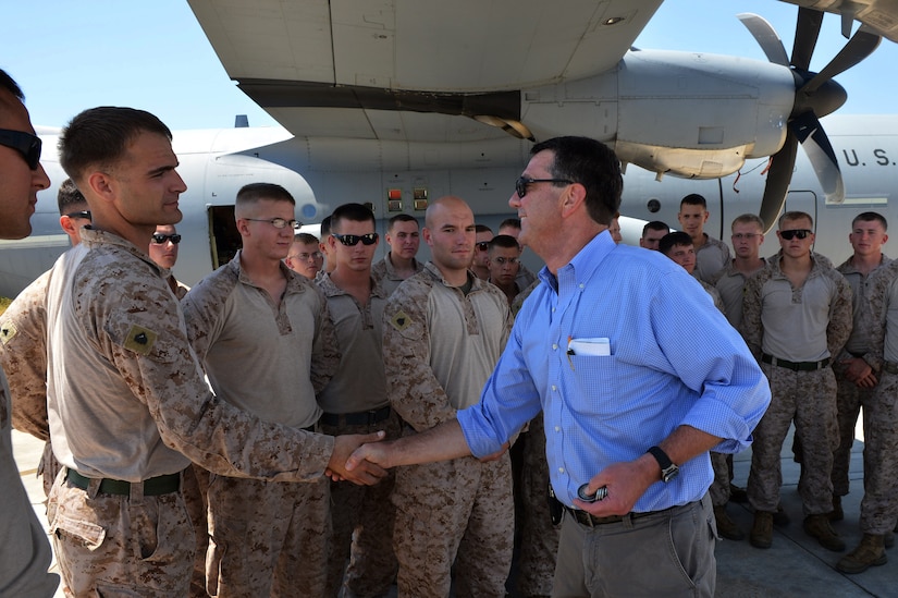 A man shakes hands with a service member as other service members stand nearby in front of an aircraft. The troops are wearing camouflage uniforms.