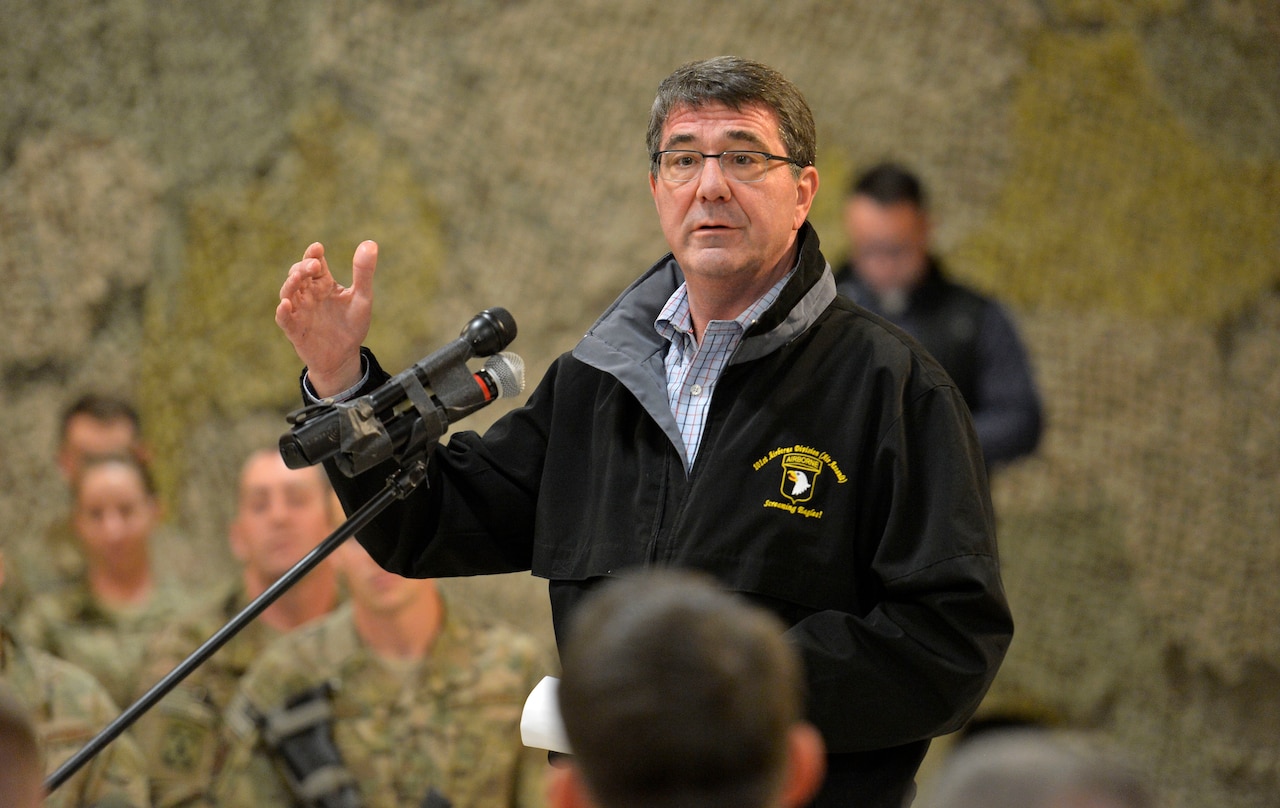 A man in a black jacket speaks from a podium to service members in camouflage uniforms. Camouflage netting hangs behind him.