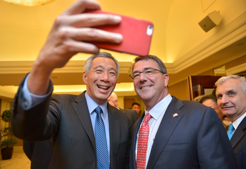 A man holds a cell phone aloft with his right hand while posing with another man for a selfie.