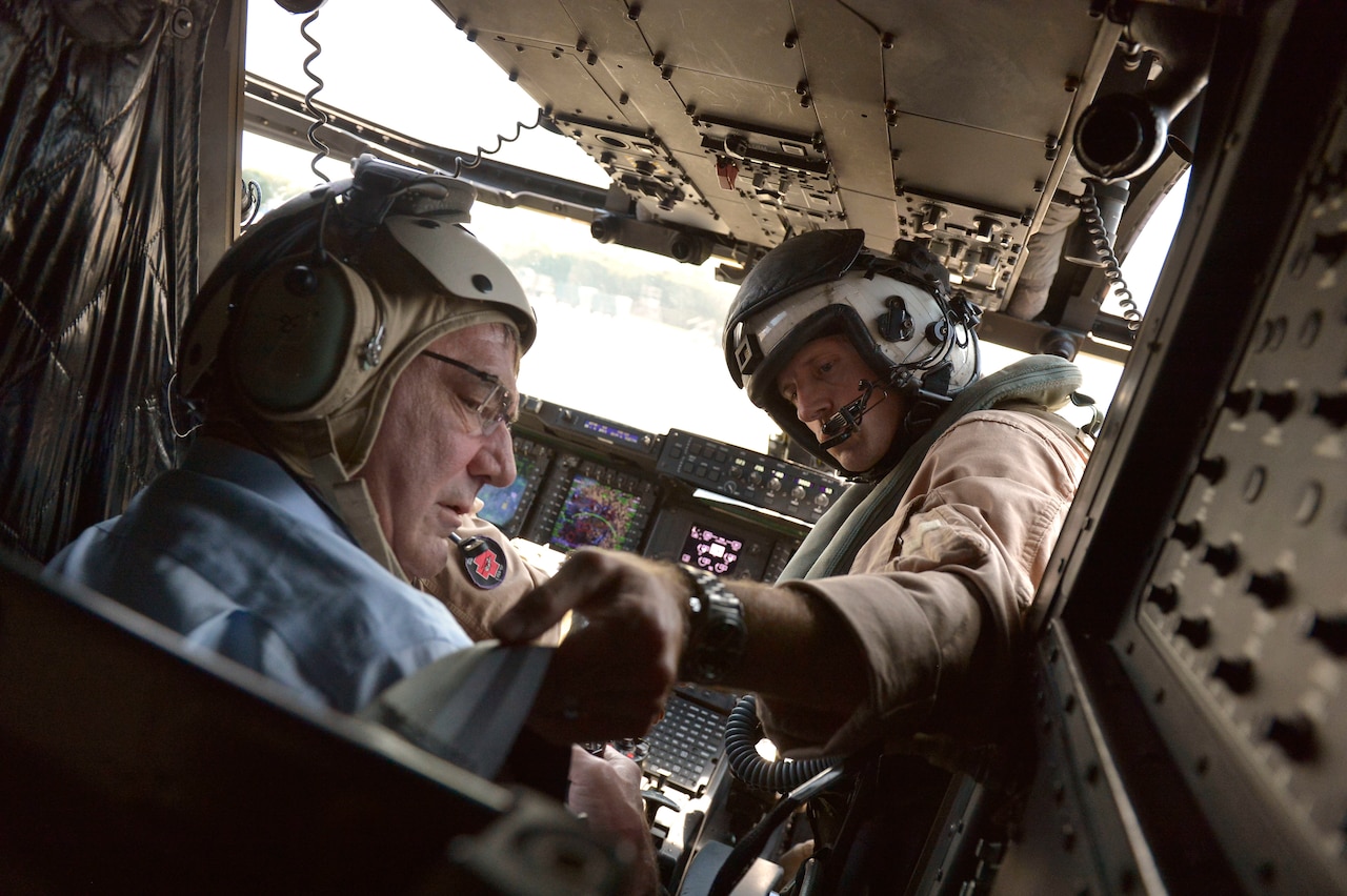 A man in a flight suit and helmet helps a second man wearing business clothes and a flight helmet with his harness straps inside an aircraft.