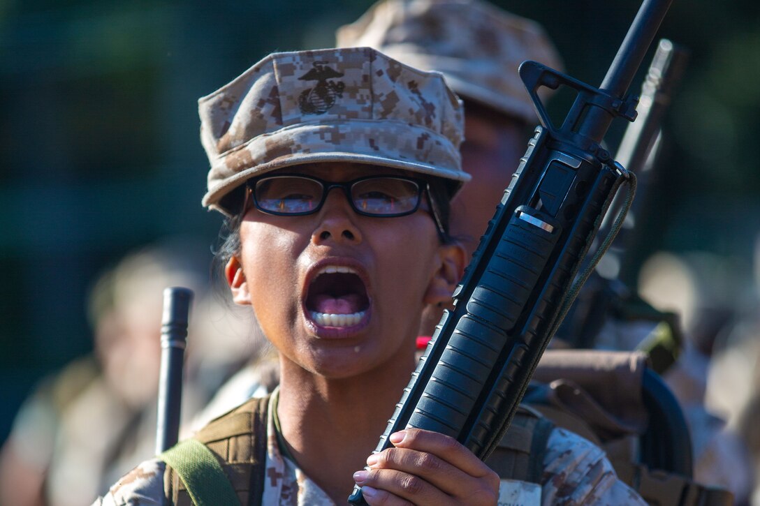 A Marine recruit leading a line of others shouts a call while holding a training rifle.