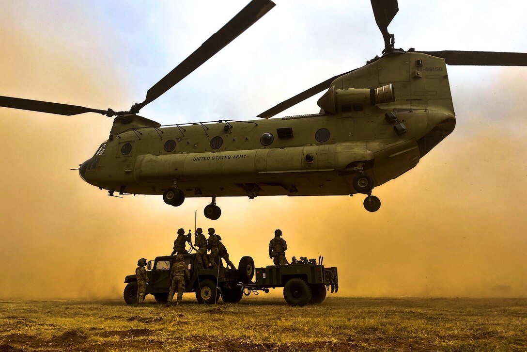 Soldiers stand on a vehicle to hook underneath a hovering Chinook aircraft surrounded by sandy dust clouds.