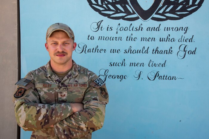 Senior Airman Kyle Riley, 332d Expeditionary Civil Engineering Squadron Explosive Ordnance Disposal Logistics Technician, in front of a morale T-wall at an undisclosed location in Southwest Asia, Oct. 14, 2022. The wall displays an EOD insignia and the quote "It is foolish and wrong to mourn the men who died. Rather we should thank God such men lived." The quote is attributed to George S. Patton. (U.S. Air Force photo by: Tech. Sgt. Jim Bentley)