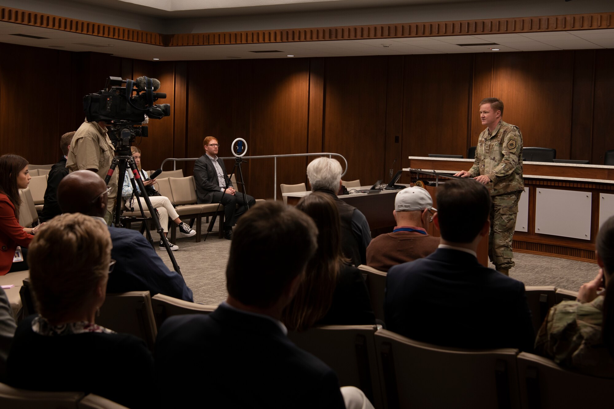 A crowd sits in a room with a military member speaking to him.