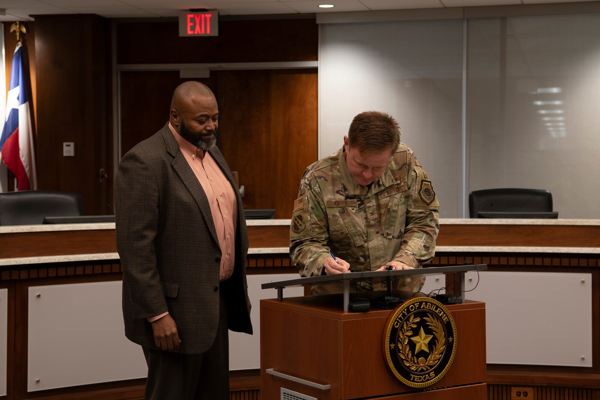 Someone signs while leaning over a podium while another watches.