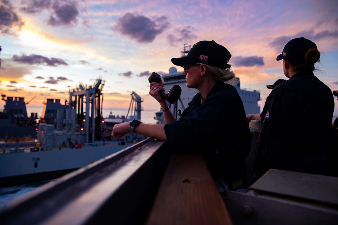 A sailor standing on the deck of a ship holds a telephone receiver at twilight.