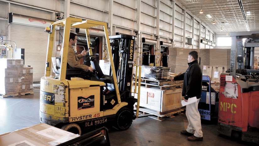 A man drives a forklift in a warehouse beside another man near a pallet of supplies.