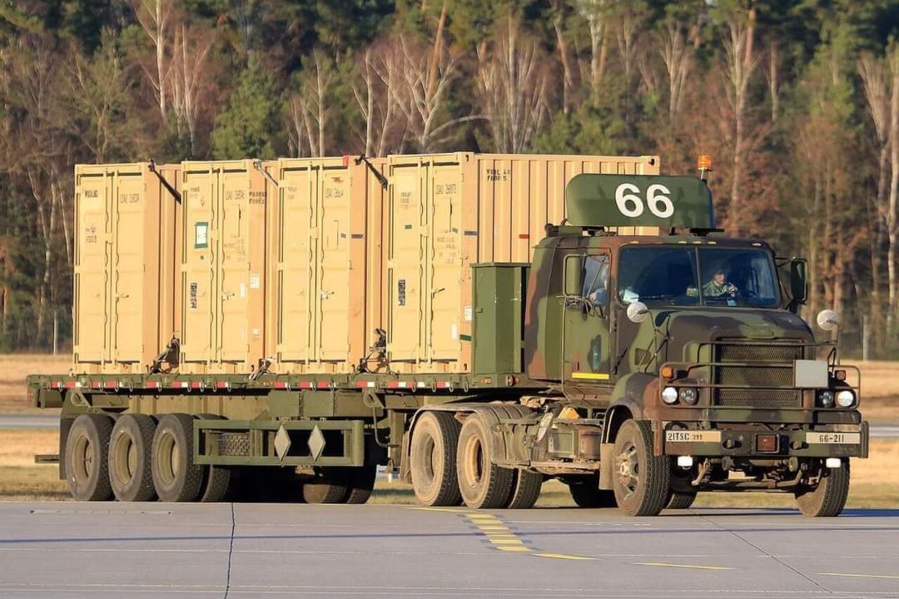 A service member drives a military truck on a road in a wooded area.