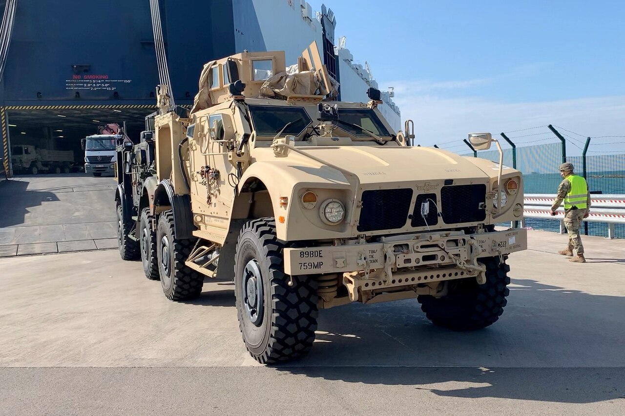 One military vehicle sits at the foot of a  ramp as another one follows behind.