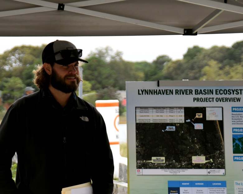 Beareded man in ball cap and sunglasses speaking and referring to an information board fo the Lynnhaven River Ecosystem Restoration Project while standing under a tent and trees in the background