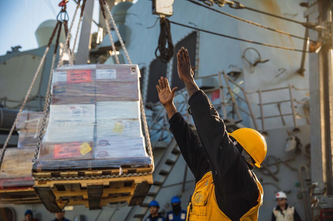 A sailor holds both arms up as a cargo of boxes hangs in the air.