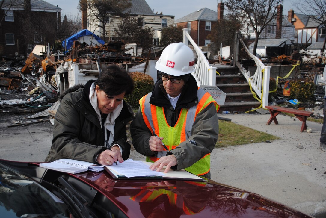 A woman signs papers on the hood of a car with another woman in a hardhat and safety vest. A burned down house appears behind them.