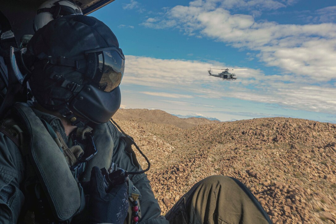 A Marine in an airborne helicopter looks at a helicopter flying nearby over mountains.
