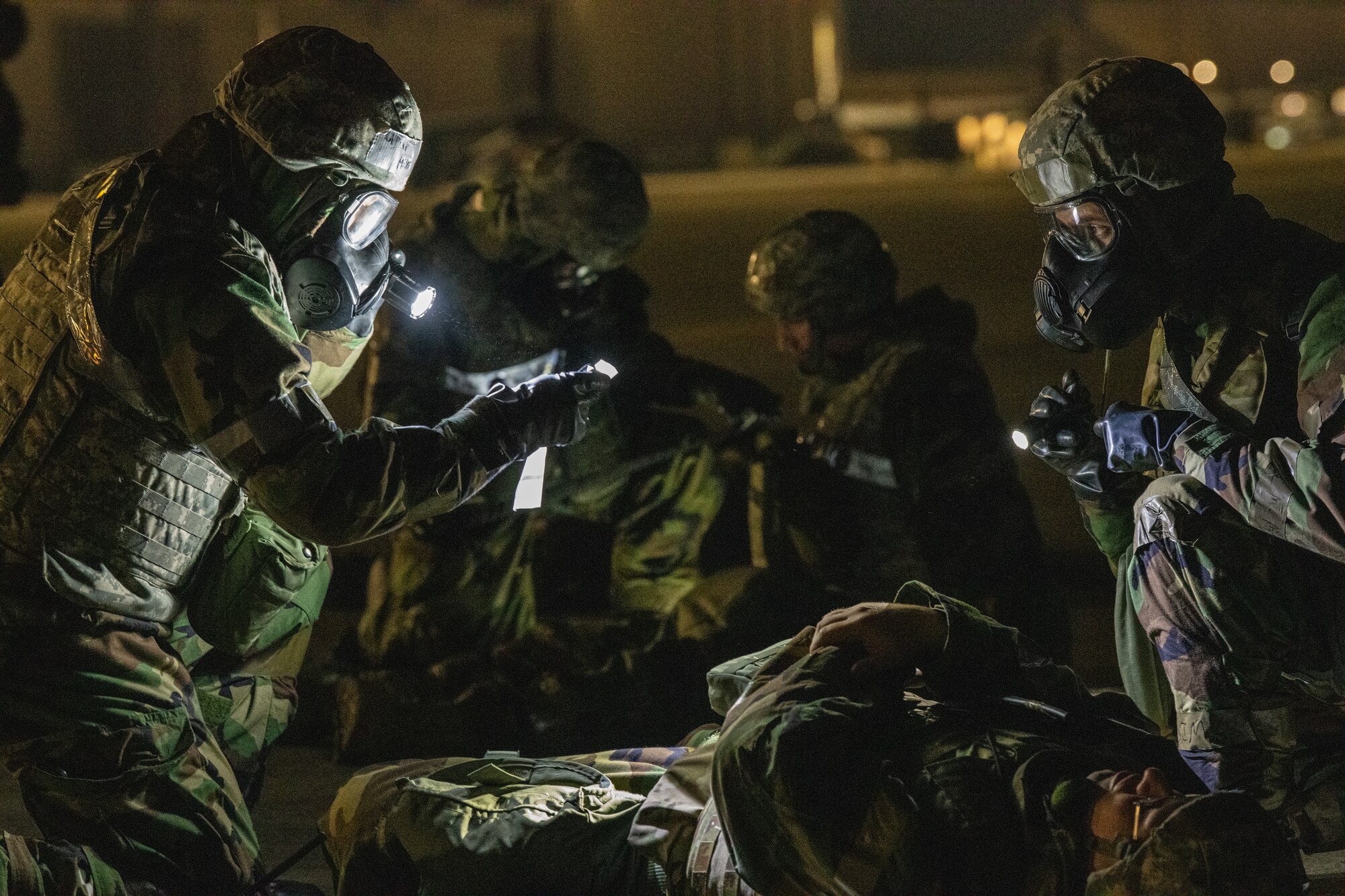 An airman in protective gear uses a flashlight to read a patient's information sheet