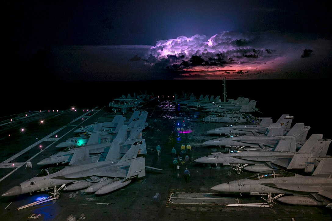 Sailors standing next to aircraft aboard a ship travel through a storm illuminated by colorful lights.