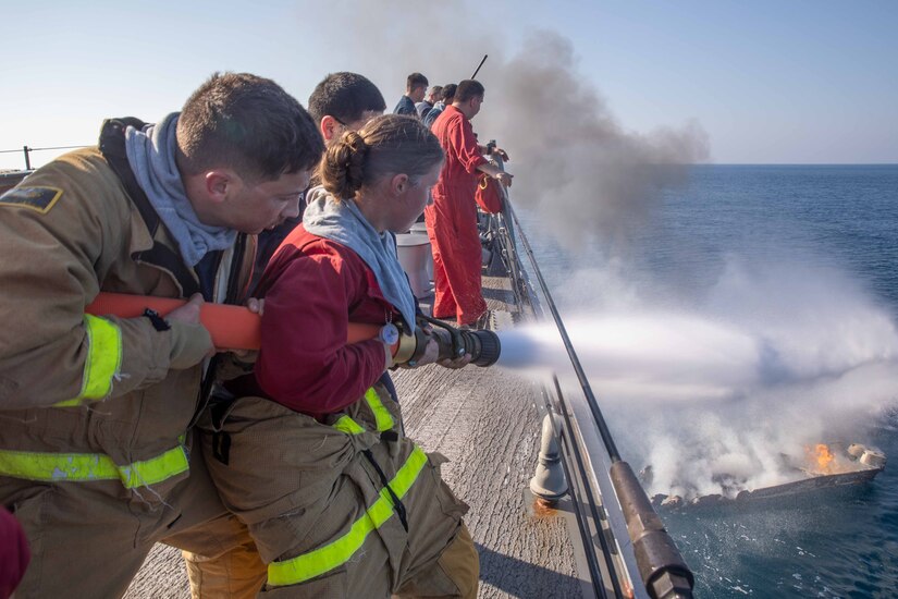 A group of sailors on the deck of a ship use water hoses to put out fire on a small boat below.