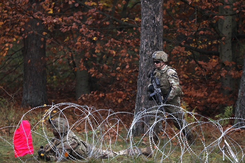 A soldier with a gun walks toward barbed wire strung across the ground in a wooded area.