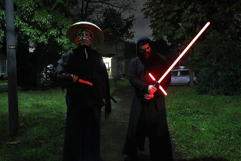 Two children in Halloween costumes with fluorescent swords stand in a yard.