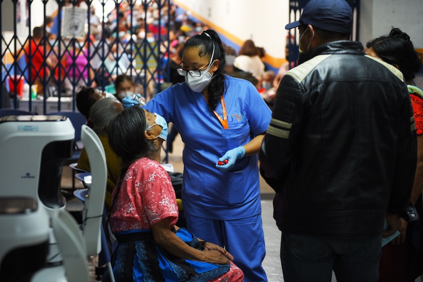 A seated woman looks upward while another woman in blue scrubs puts drops into her eyes.
