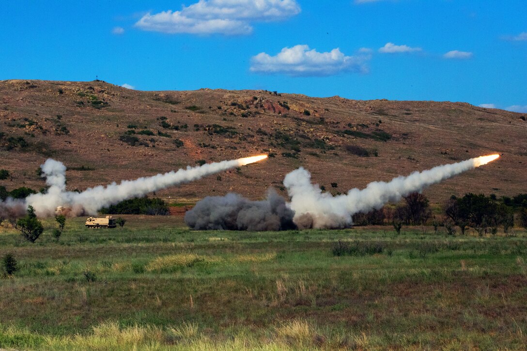 Two rockets launch in a field, creating parallel lines of smoke.