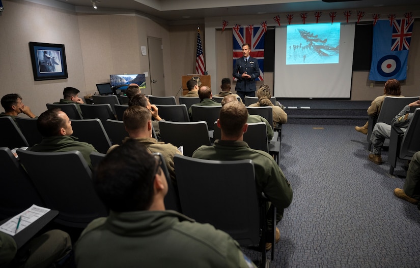 Squadron Leader Maj. David Wild, 94th Fighter Squadron pilot, briefs participants on the Battle of Britain in the 94th FS briefing room at Joint Base Langley-Eustis, Virginia, Oct. 21, 2022.  Wild, a British Royal Air Force pilot is part of the Foreign Exchange Officer Program which is a way for partner nations to strengthen alliances and share organizational knowledge to further their home’s lethality. The  94th FS is the only F-22 squadron in the Air Force with a British FEO. (U.S. Air Force photo by Tech. Sgt. Matthew Coleman-Foster)