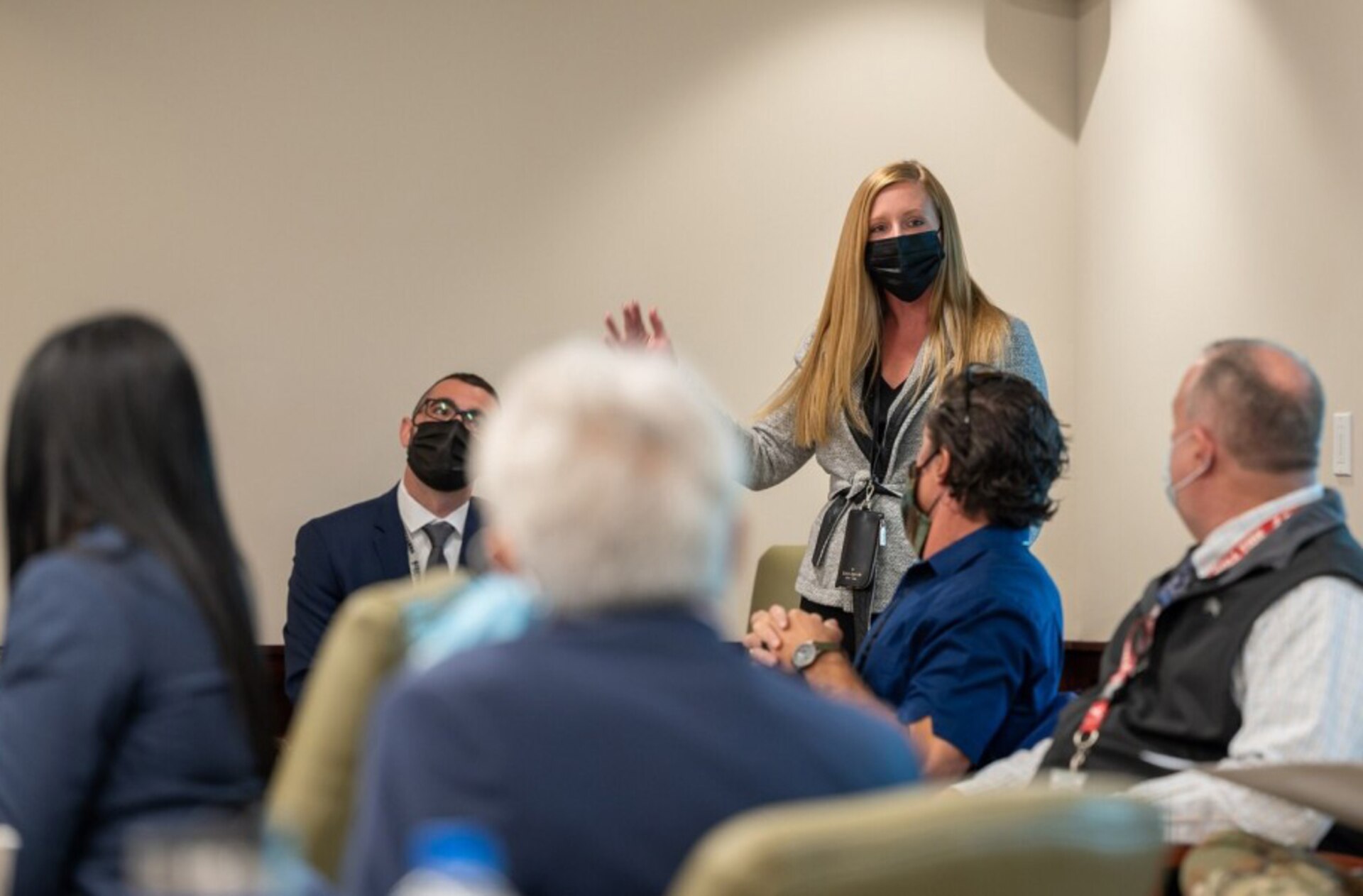 Photo of a women stands in a room to give a briefing before a group of people seated around a table.
