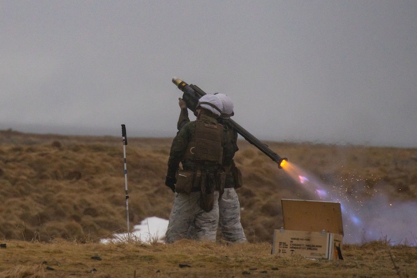 Two service members stand in a field and fire a shoulder-launched missile.