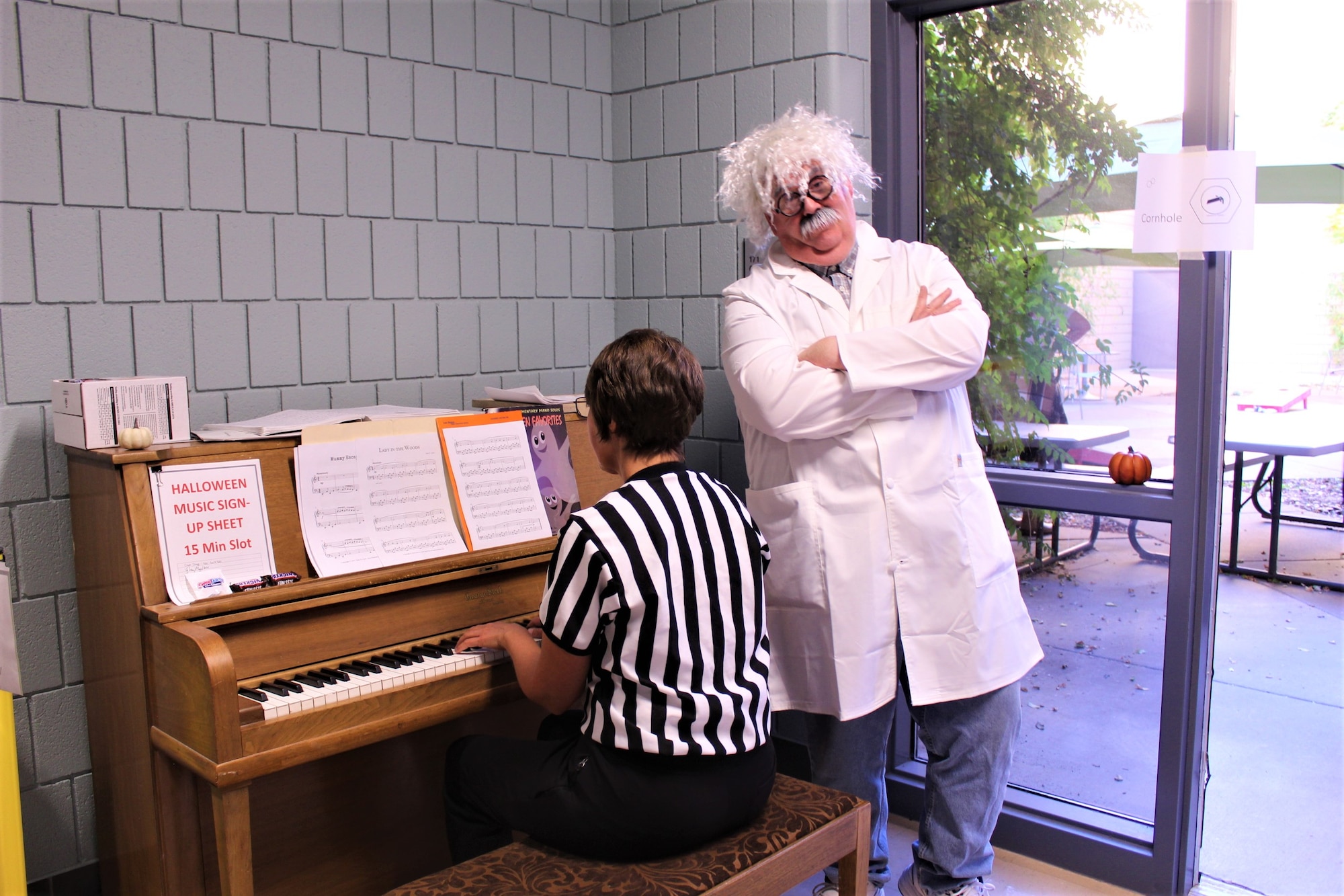lady plays piano while a man stands nearby in a scientist costume