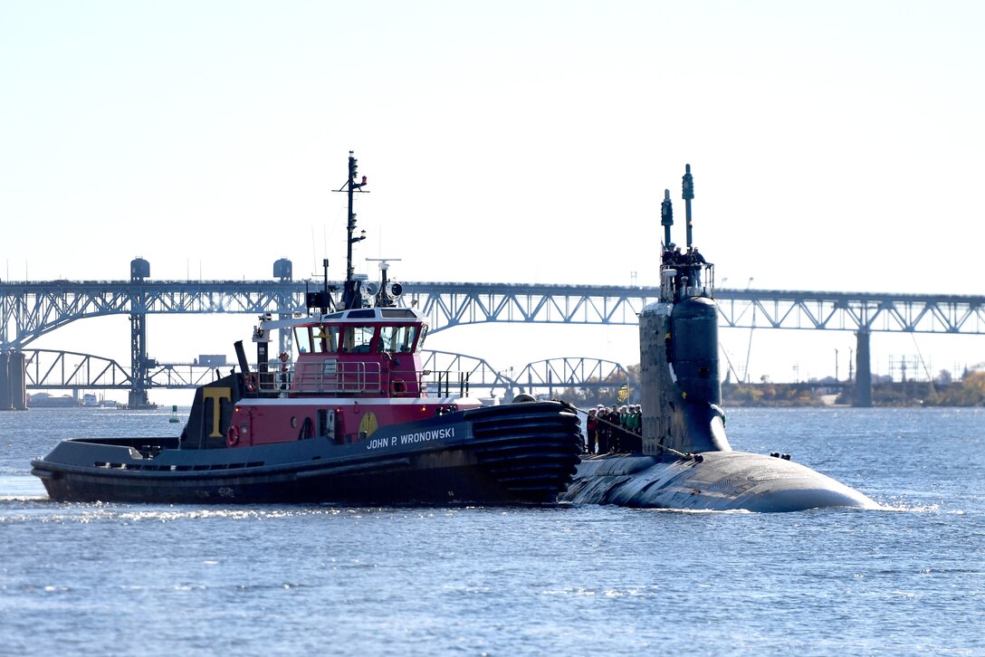 A Navy submarine travels with a tugboat in a river, with a bridge in the background.
