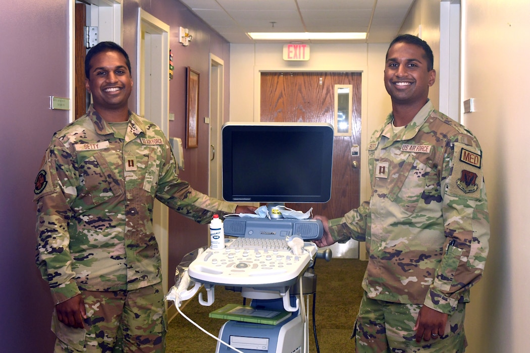 Identical twins in camouflage uniforms stand on either side of an ultrasound machine