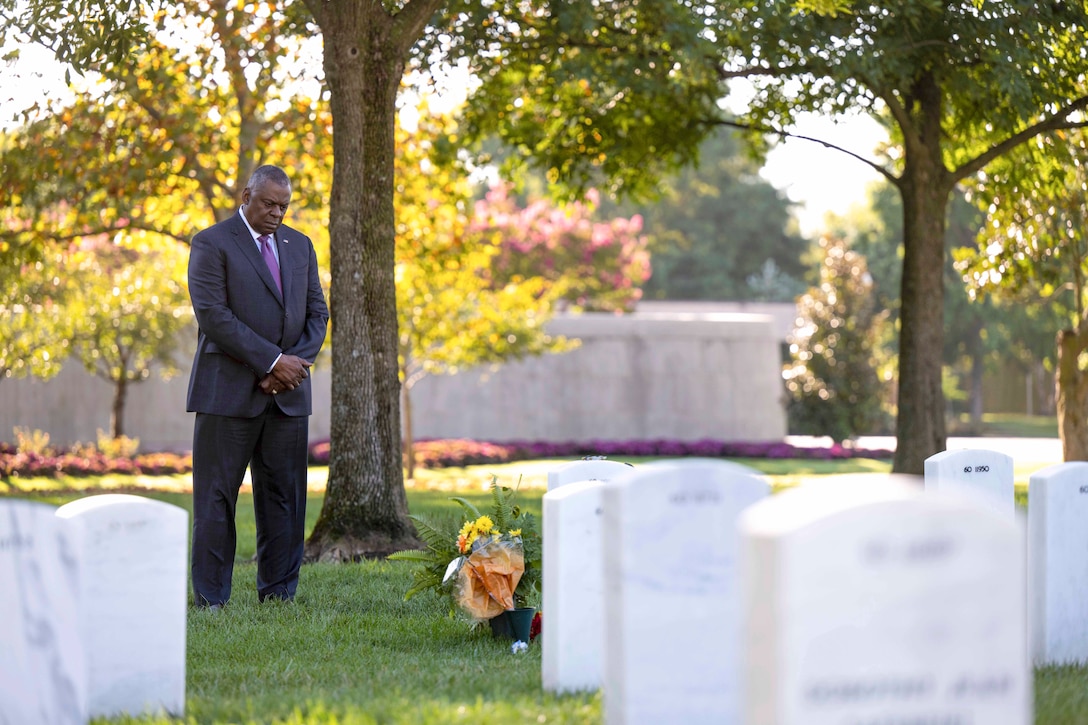 The secretary of defense looks down while standing in a cemetery.