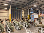 A man in camo uniform addresses men and women also wearing camo sitting in metal folding chairs in a warehouse.