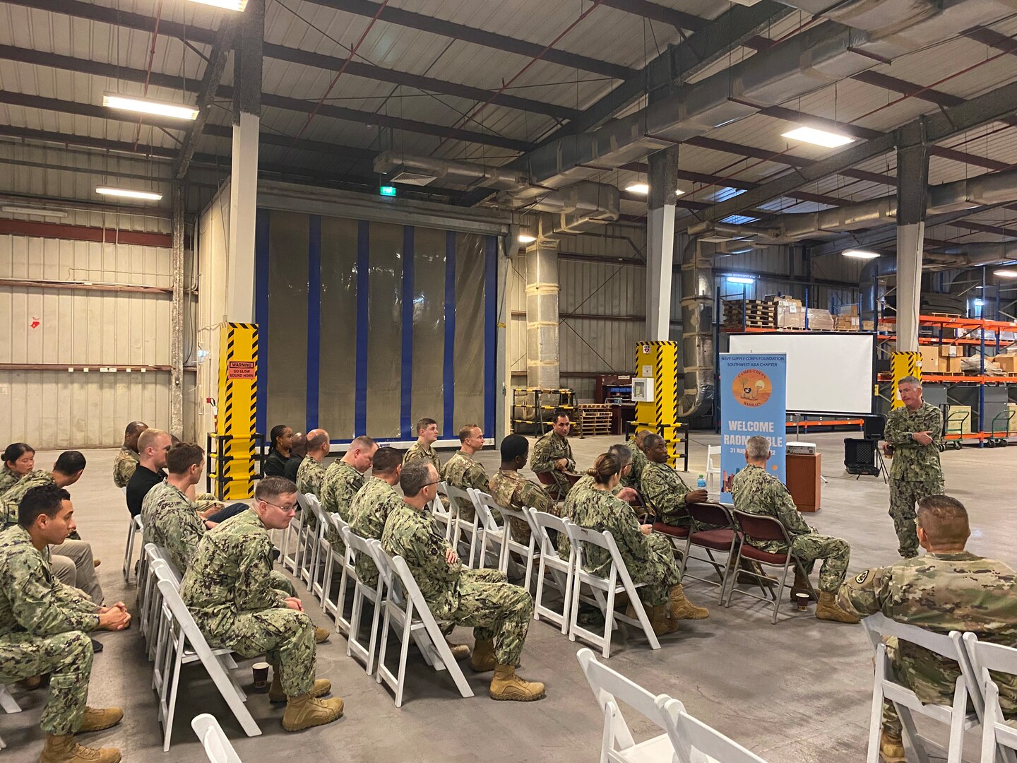 A man in camo uniform addresses men and women also wearing camo sitting in metal folding chairs in a warehouse.